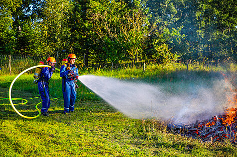 Zwei Feuerwehrleute (vermutlich Jugendfeuerwehr) in blauen Anzügen und orangefarbenen Helmen löschen mit einem Feuerwehrschlauch ein kleines Flammen- und Rauchfeuer auf einer Wiese, im Hintergrund Bäume und sommerliche Vegetation.