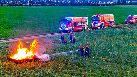 Jugendfeuerwehrmitglieder in Schutzkleidung und orangefarbenen Helmen bekämpfen auf einer Blumenwiese mit einem Schlauch ein offenes Übungsfeuer in einer rechteckigen Metallwanne. Im Hintergrund stehen mehrere Feuerwehrfahrzeuge auf einem Feldweg, während weitere Jugendliche das Geschehen beobachten oder unterstützen. Am Horizont ist eine Siedlung zu sehen.