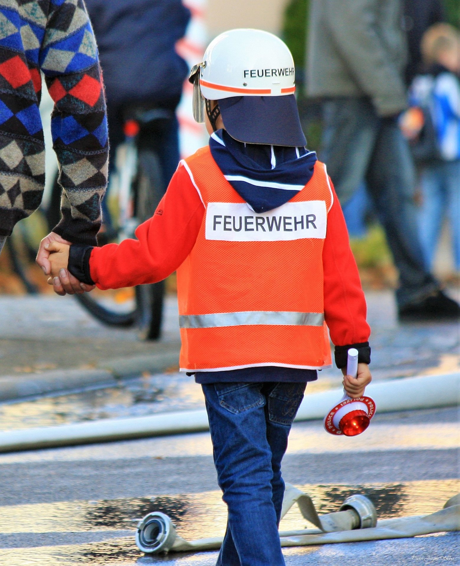 Kleiner Junge in Feuerwehrkleidung an der Hand eines Erwachsenen