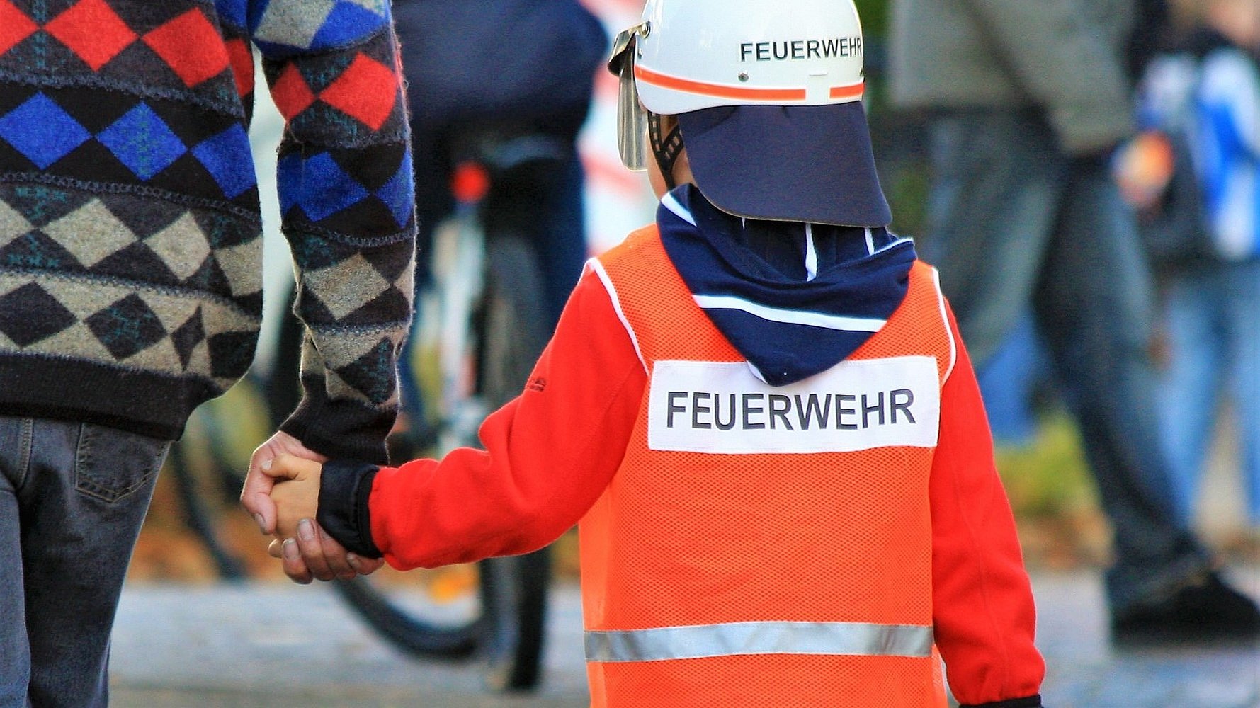 Kleiner Junge in Feuerwehrkleidung an der Hand eines Erwachsenen