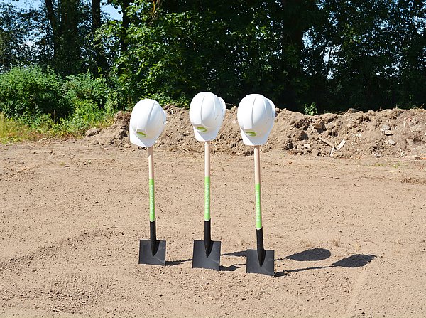 Drei Spaten stecken in der Erde. Auf jeden wurde ein Bauhelm gehängt.