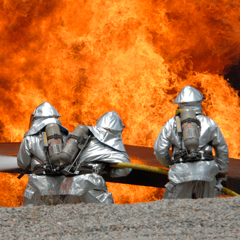 Drei Feuerwehrleute stehen in Schutzkleidung vor einem großen Feuer.